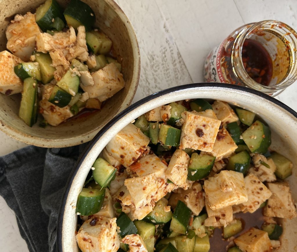 An easy spicy, Asian-style cucumber salad recipe with soft tofu, cucumbers, chili crisp/chili crunch, sesame seeds in a large bowl. A jar of chili crunch is off to the side.